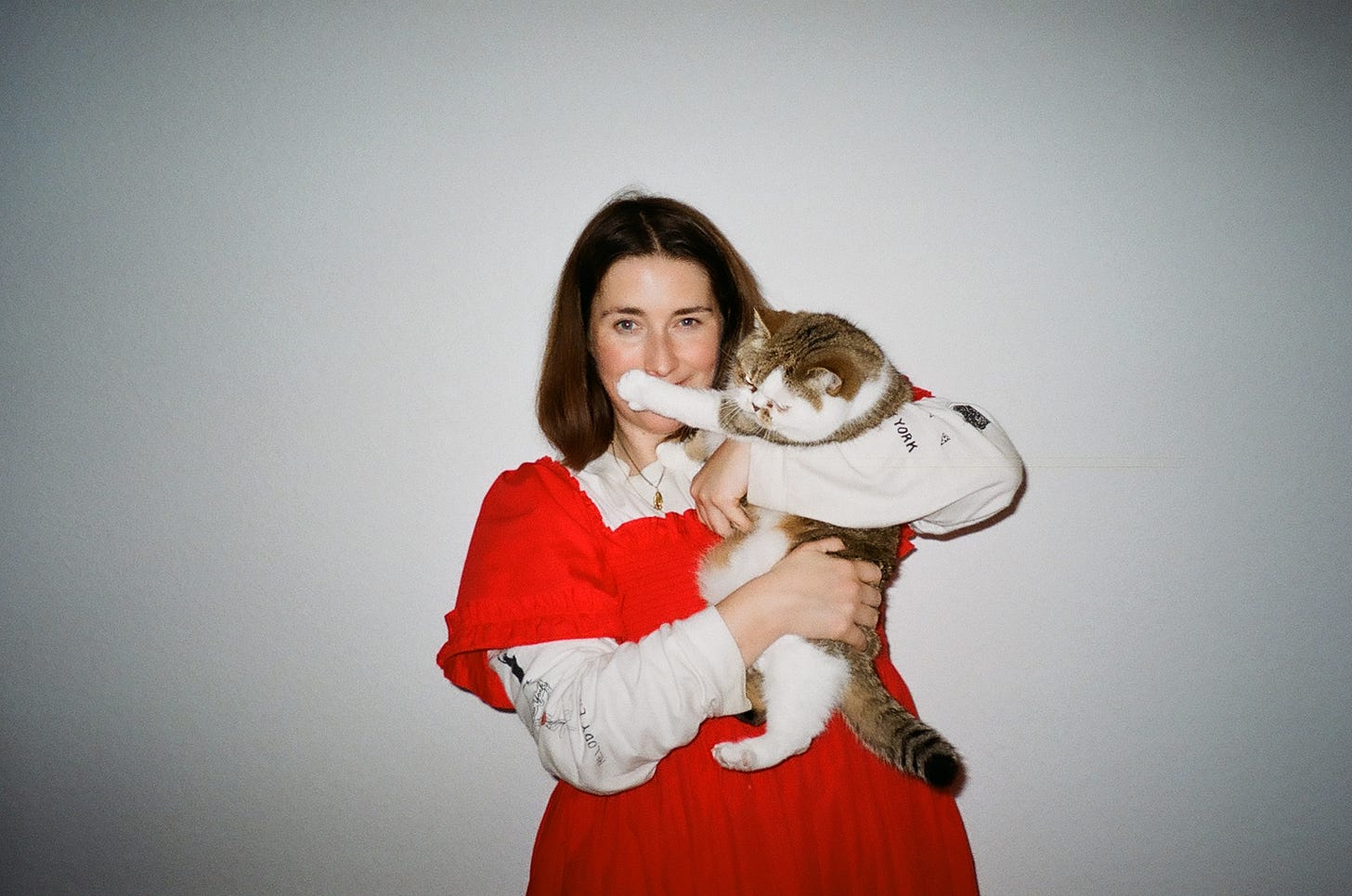 Fiona is a white woman with straight brown hair to her shoulders. In this photograph, she's standing in front of a plain white wall, wearing a bright read short-sleeved dress and a white undershirt. She's holding a cat with white and tabby-coloured fur in her arms, and the cat's paw is slightly covering her face.