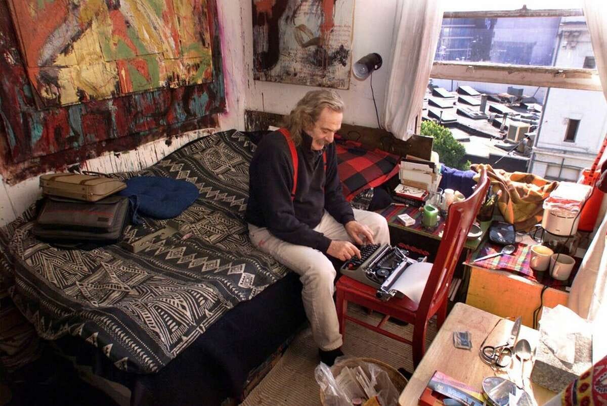 Jack Hirschman sits on the edge of his bed in his apartment. In front of him is a chair with a typewriter on it that Jack is writing on.