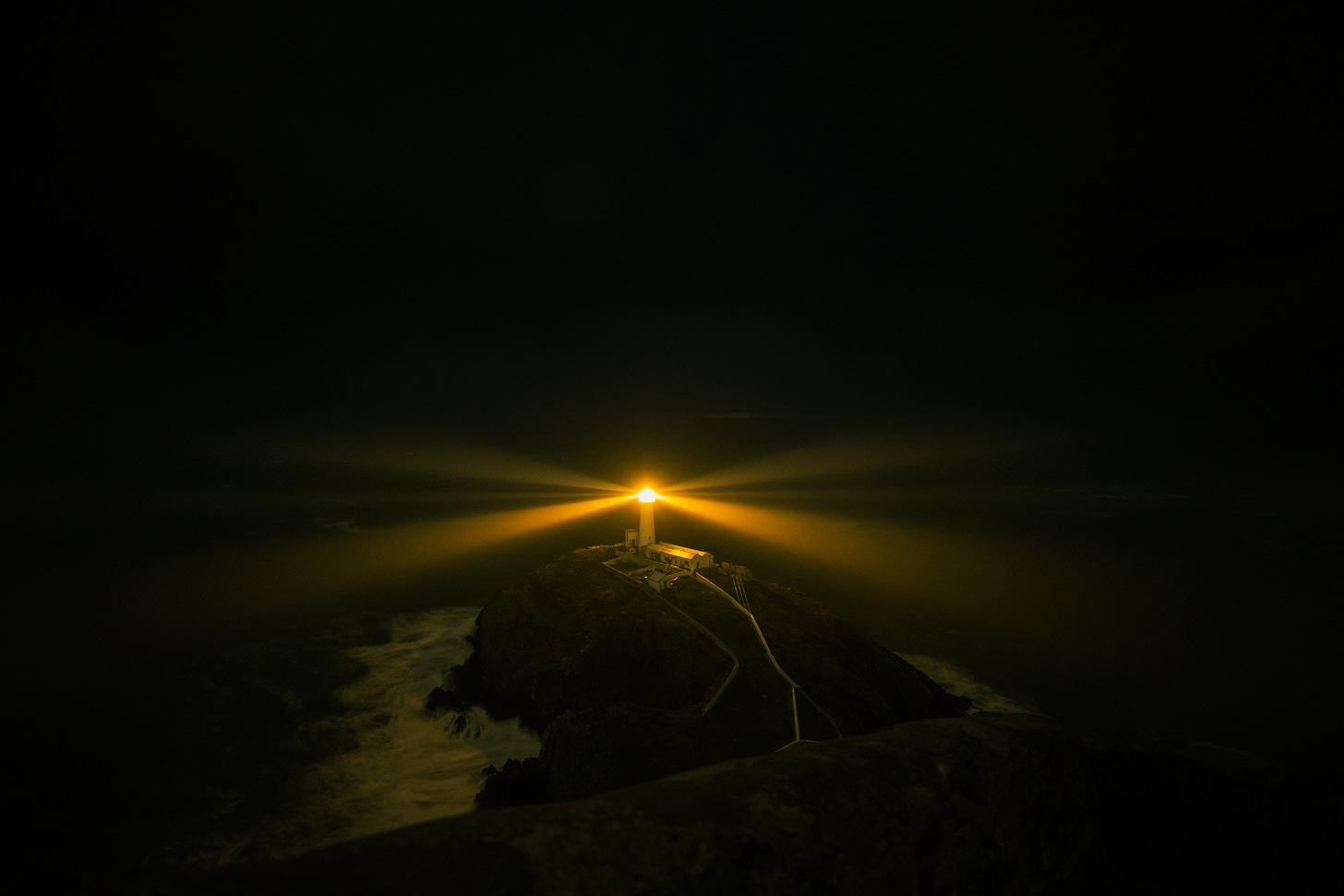 A lighthouse on the top of a hill shining orange light. The rest of the picture is black.