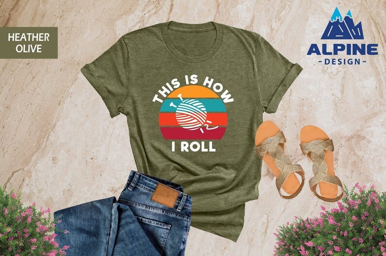 May include: A heather olive green t-shirt with a retro sunset graphic and the text 'This is how I roll' and a yarn ball with knitting needles. The t-shirt is displayed on a light brown textured surface with blue jeans and gold sandals.