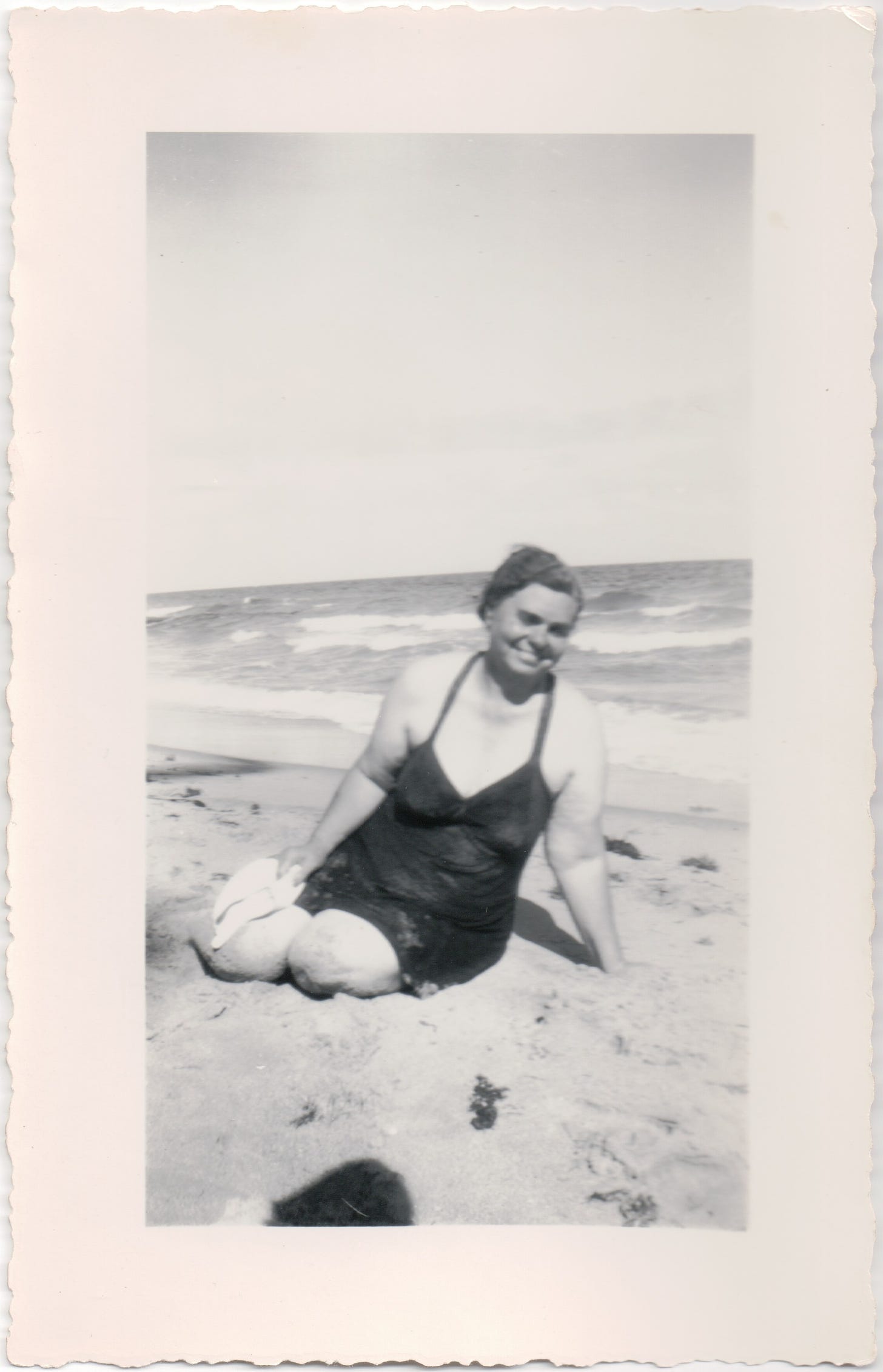 Anne Cheff Marquis, a large woman sitting on a beach wearing a bathing suit, with a big smile on her face, leaning on one arm 