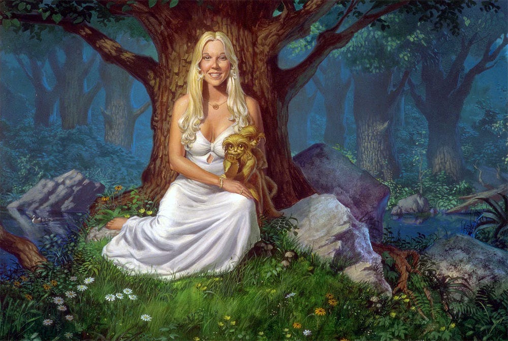 A smiling blonde woman in white dress sits on a bed of grass with her back against the wide trunk of a tree. Her knees are folded under her. She rests her right arm across to her opposite leg where a small tawny furred creature with golden eyes leans on its elbow, also looking directly at the audience. Another fuzzy stands behind it looking off. Large boulders surround the tree and a brook winds past behind them.