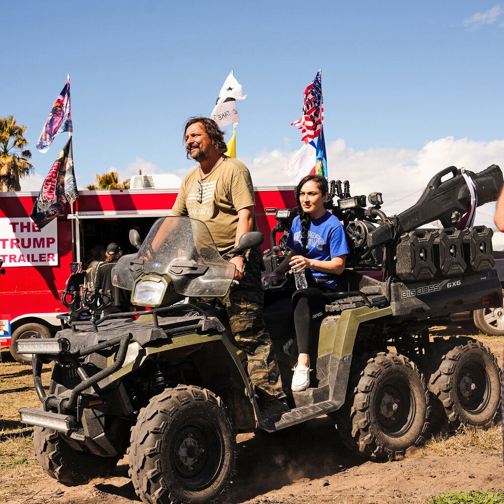 Two people ride an all-terrain vehicle amid a gathering of other people and flying flags.