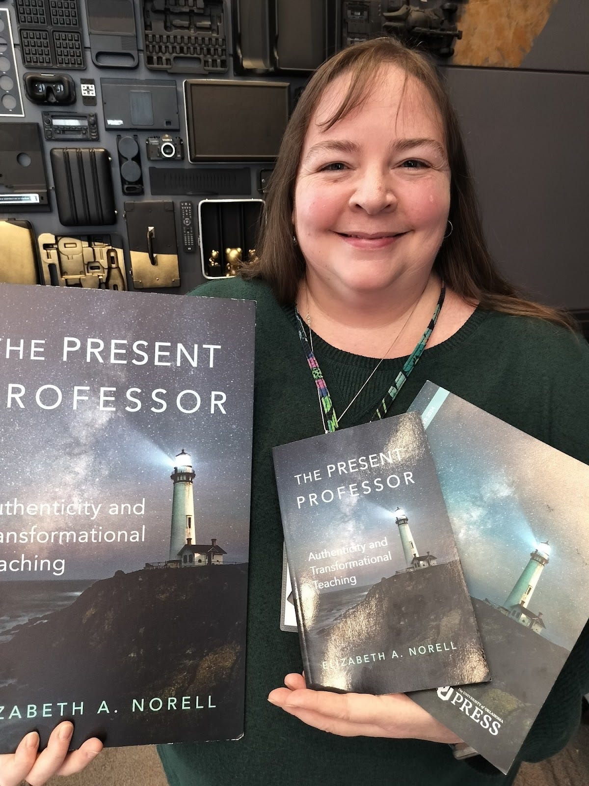 Liz holding copies of her book, a large poster, and the OU Press catalogue, all featuring an image of a lighthouse against a starry sky and the book title "The Present Professor."