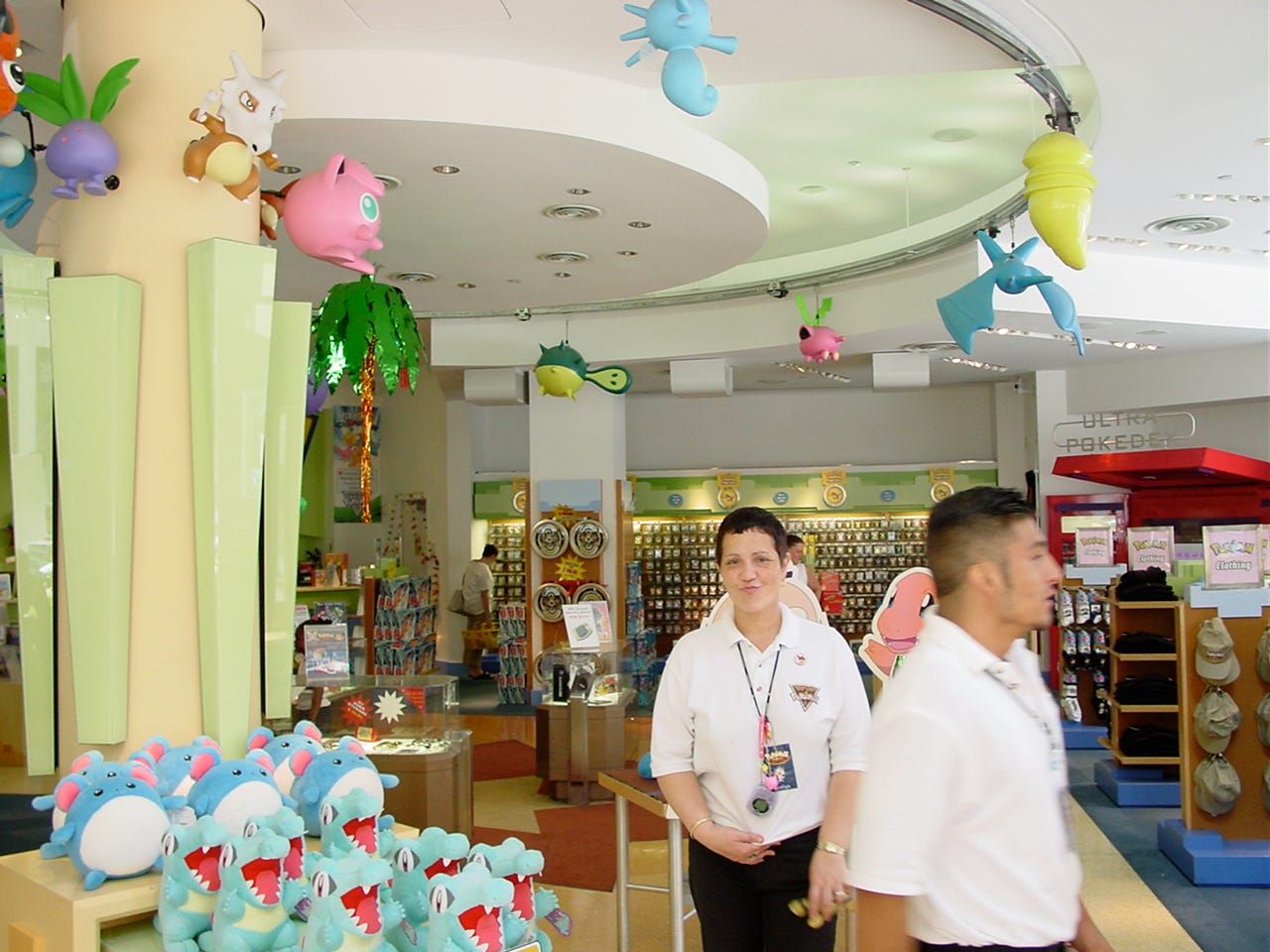 A photograph of the lower floor, showing more Pokémon animatronics, including Horsea, Kakuna, Zubat, Hoppip, Qwilfish, and Koffing. Statues such as Cubone, Jigglypuff, and Eevee can also be seen. A huge range of merchandise, and Pokémon Center staff, are also present in the photograph