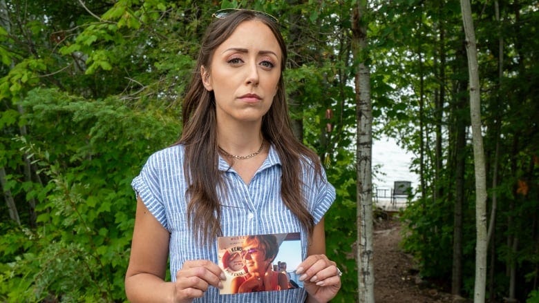 A young woman stands in front of trees, holding a photograph.