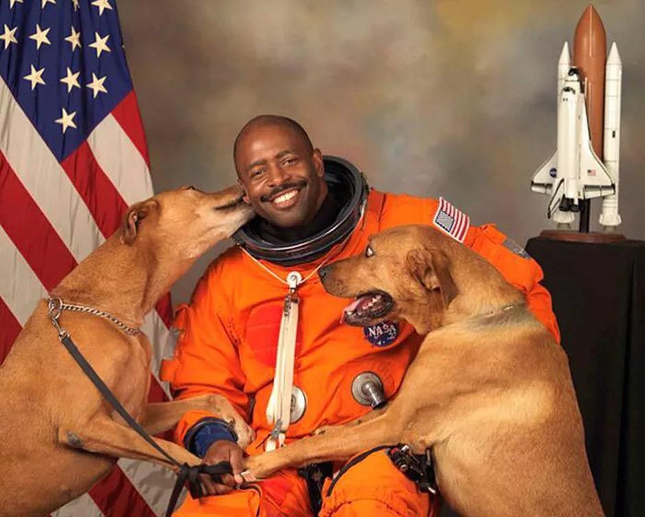 Astronaut Leland Melvin in an orange space suit, no helmet, with his two large happy dogs. He's smiling, there's a US flag and a model shuttle