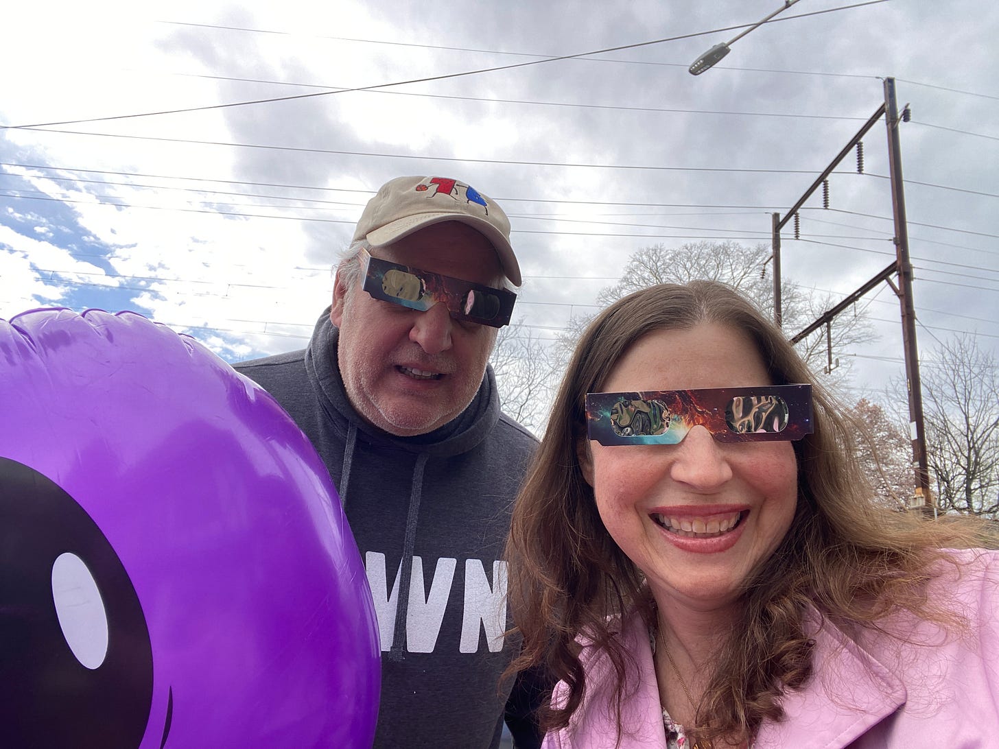 Fred and Gabrielle wear sclipse glasses and smile at the camera. Behind them  is a cloudy, overcast sky.