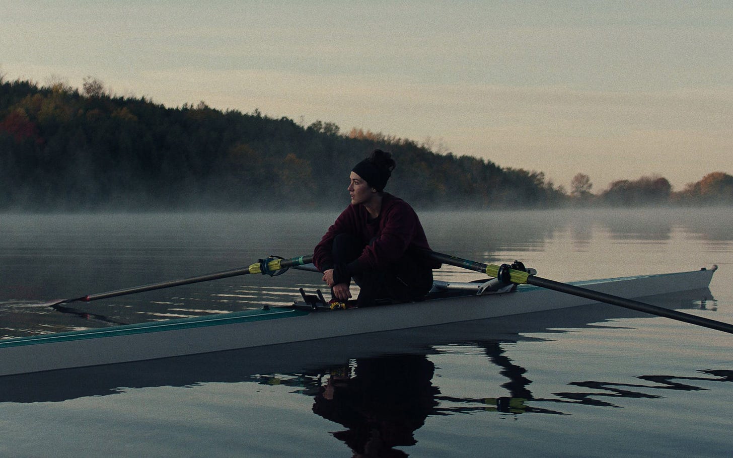 A white woman looks off into the distance as she sits with her arms draped over her legs in a rowing scull on a misty lake in the early morning.