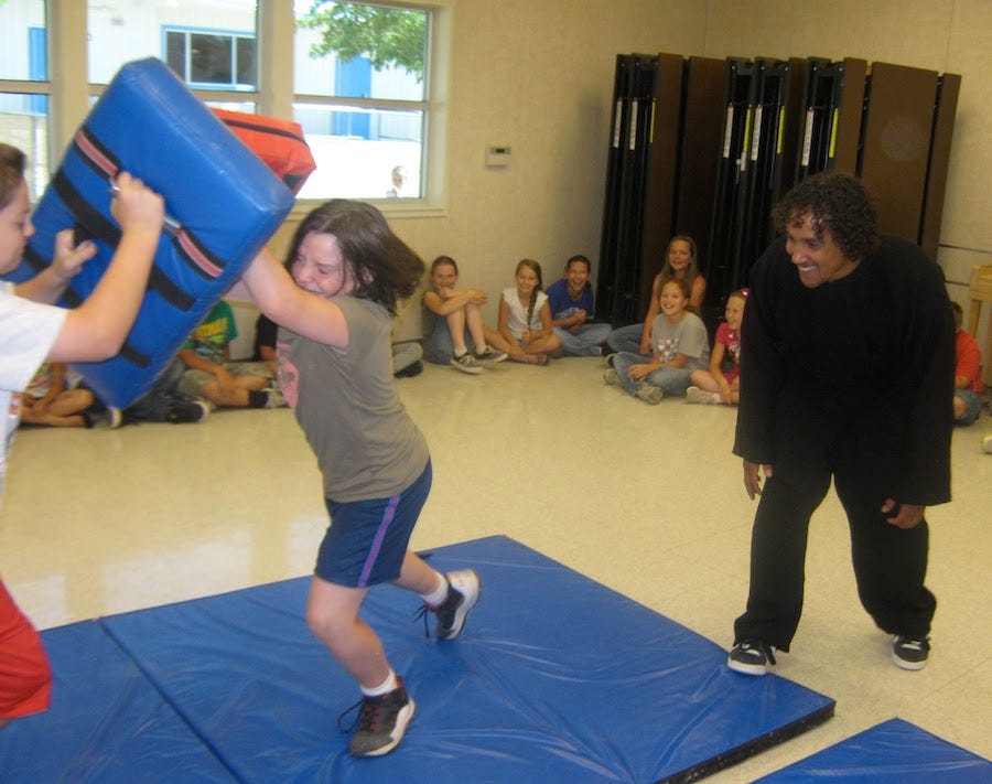 A group of children watching a Tai Chi teacher supervise a boy and a girl using large motor control to block each other with large padded blocks.