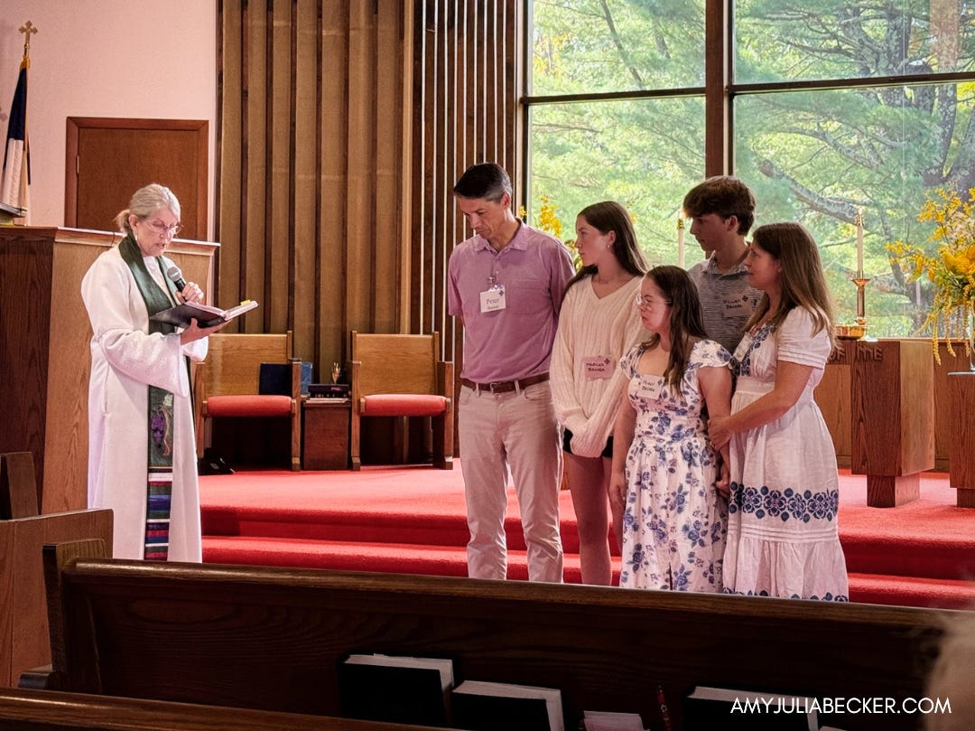 Amy Julia's family stands at the front of the church facing the congregation. The pastor stands next to them reading