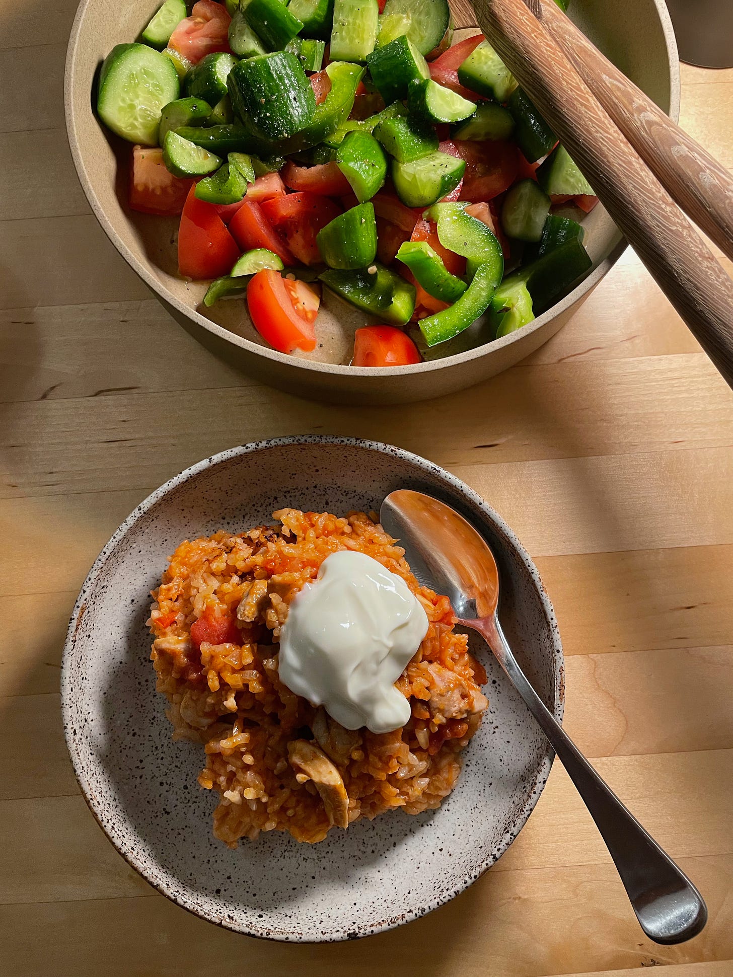 A bowl with chicken and tomato rice topped with yoghurt. A Greek-style salad with cucumber and tomato in the background.