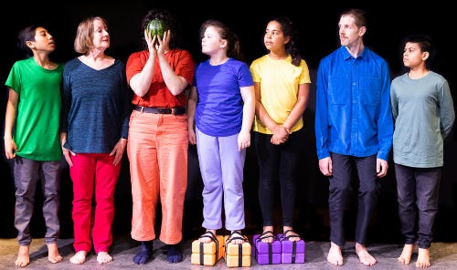 a group of children and adults in colourful clothing, lined up. One is holding a watermelon in front of her face. Some people have yoga blocks strapped to their feet so everyone is the same height