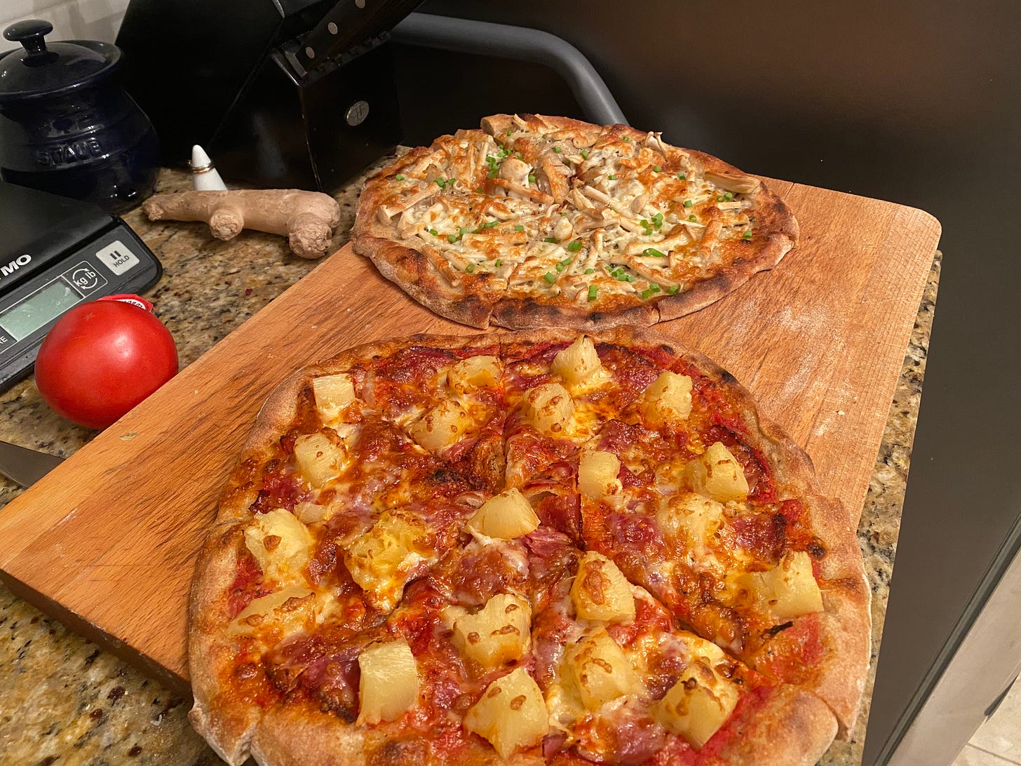 The two pizzas described above, on a cutting board on the counter: capocollo pineapple in the foreground, and mushroom in the back, topped with sliced green onions. Both pizzas have browned, bubbled cheese and browned crusts.