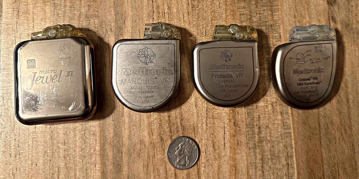 On a blonde wood table four heart defibrillators of various vintages are lined up above a quarter coin for scale.