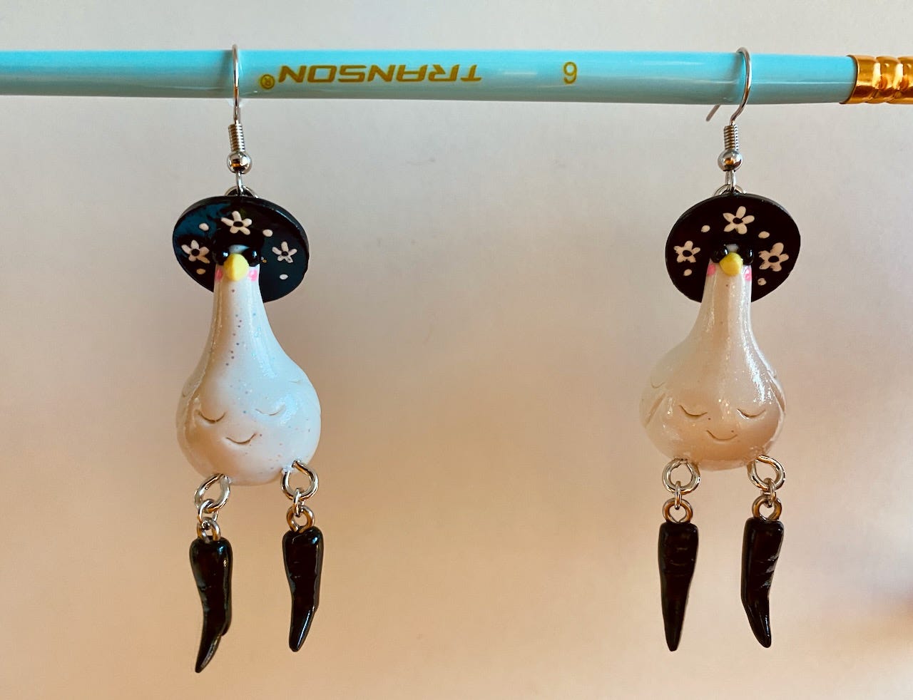 A pair of earrings hanging like fruit from a paintbrush branch. The earrings are glittery polymer clay geese wearing floral witch hats, sunglasses, and kicky black boots.