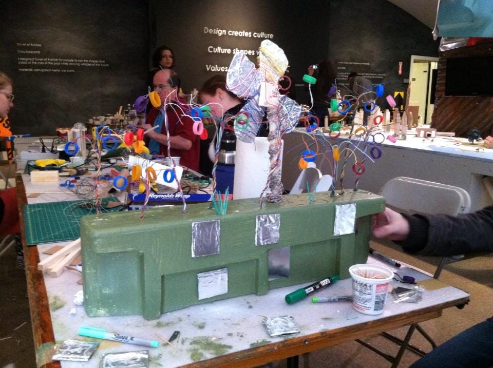 People building models at a tabel covered in art and craft supplies. In the foreground is a green foam structure with aluminum foil windows and topped by a paper wind turbine and pipe cleaner trees.