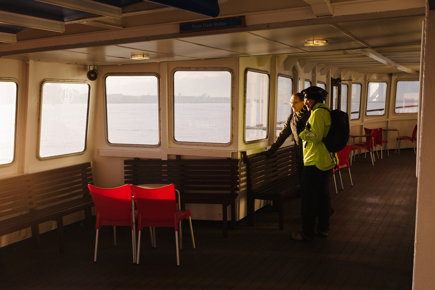 Two men stand talking inside the top deck of the ferry on an early morning ride to Liverpool. One man is wearing a hi-vis green jacket and there are red plastic chairs glowing in the early light.
