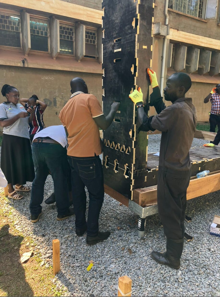 2 people hold up a WikiHouse wall block made of timber and painted black. Another person helps them attach the block to the floor.