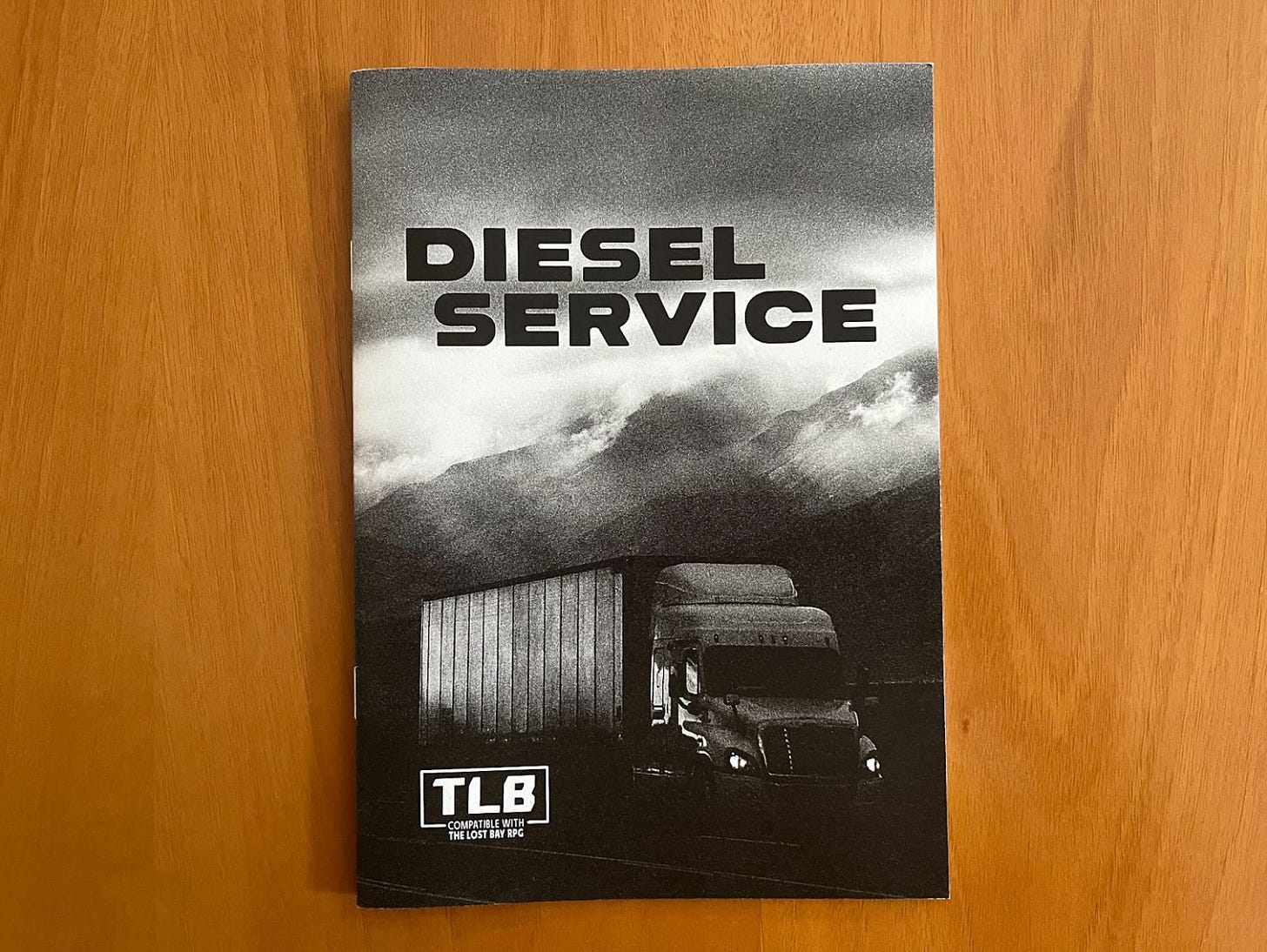 The zine Diesel Service, designed by Allen Hall, compatible with the Lost Bay. The cover is black and white and features a semi-trailer truck on a road in front of a mountain. The sky is overcast, with sunbeams breaking through the clouds.