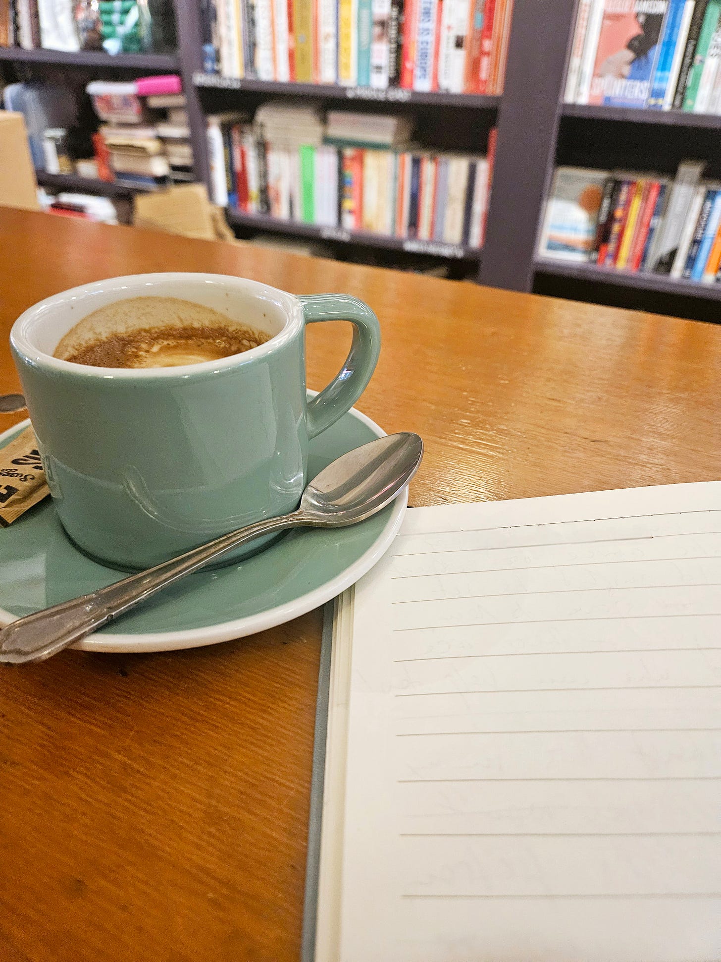 a green coffee cup is placed next to a notebook on a wooden table, and in the background, there are shelves with books.