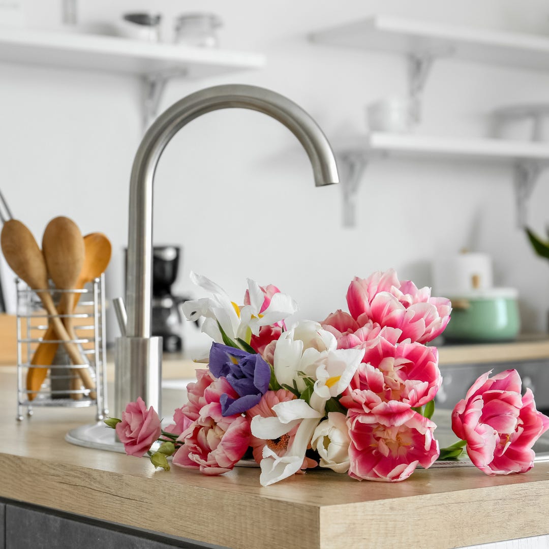 Close up of pink, white, and purple flowers in a kitchen sink. The rest of the kitchen is blurred in the background, but you can make out wooden spoons, a coffee machine, and some white shelves on the wall.