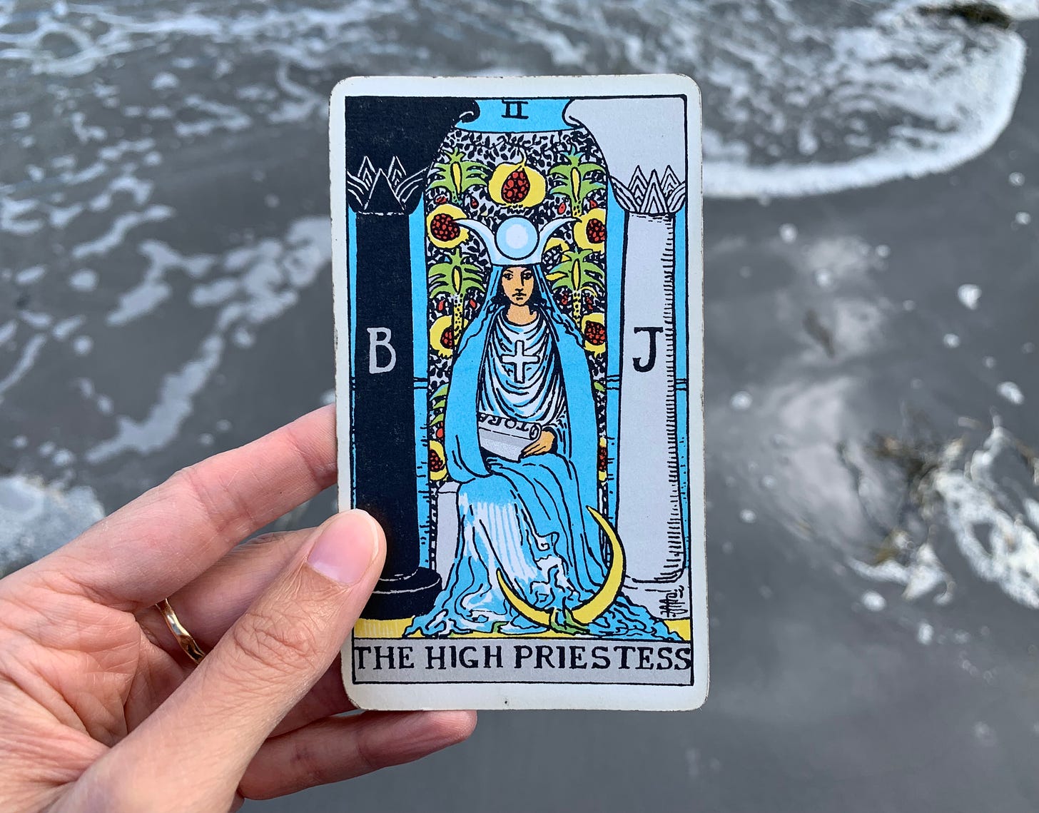 A hand is holding a tarot card, High Priestess by Pamela Colman Smith. In the card, a person who appears to be wearing a robe and veil made of water is sitting in front of a body of water that is veiled by a pomegranate curtain. She is holding a scroll. The roman numeral number two is up top. There are two pillars on either side of her, one black and one white. Behind the card is ocean water that has just rolled up and is on its descent back to the ocean.