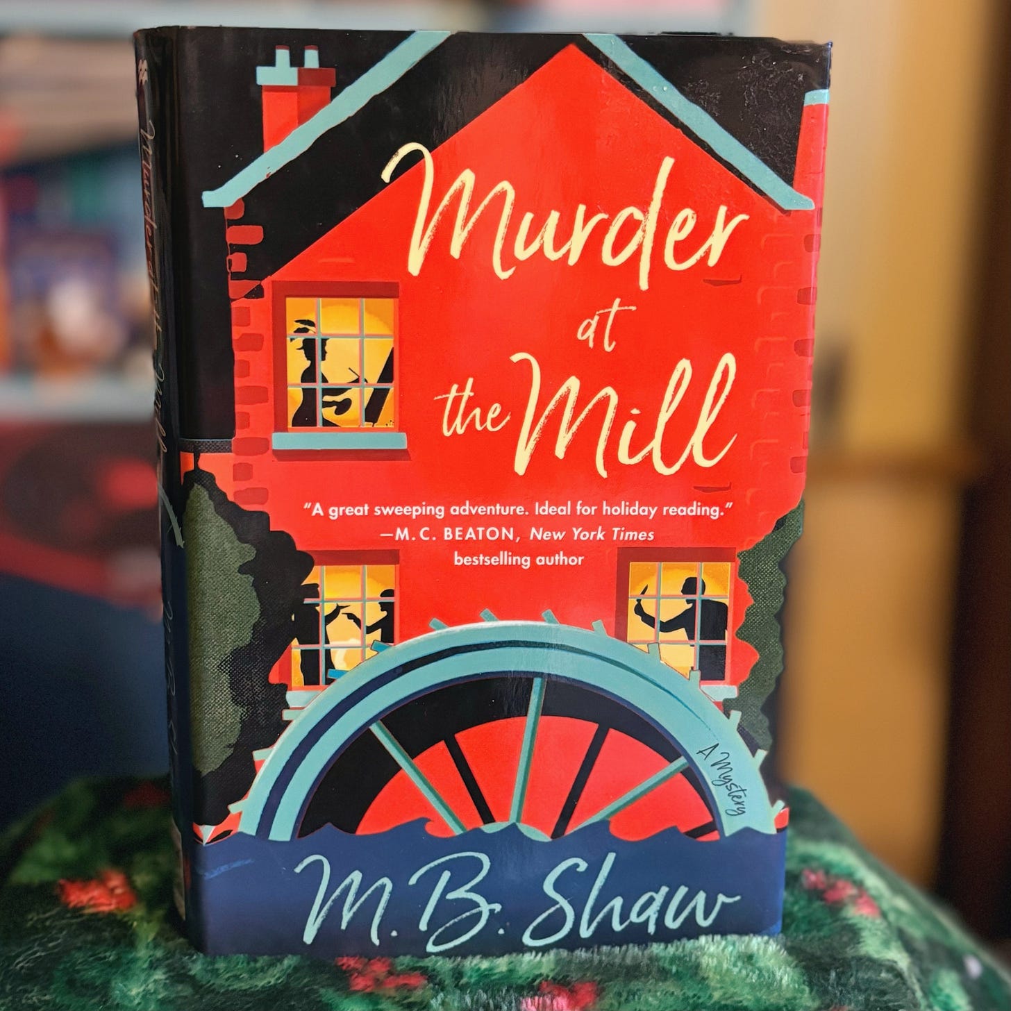 Book cover of Murder at the Mill by M.B. Shaw. Depicts at the side view of a red mill with three windows and shadows of people. 