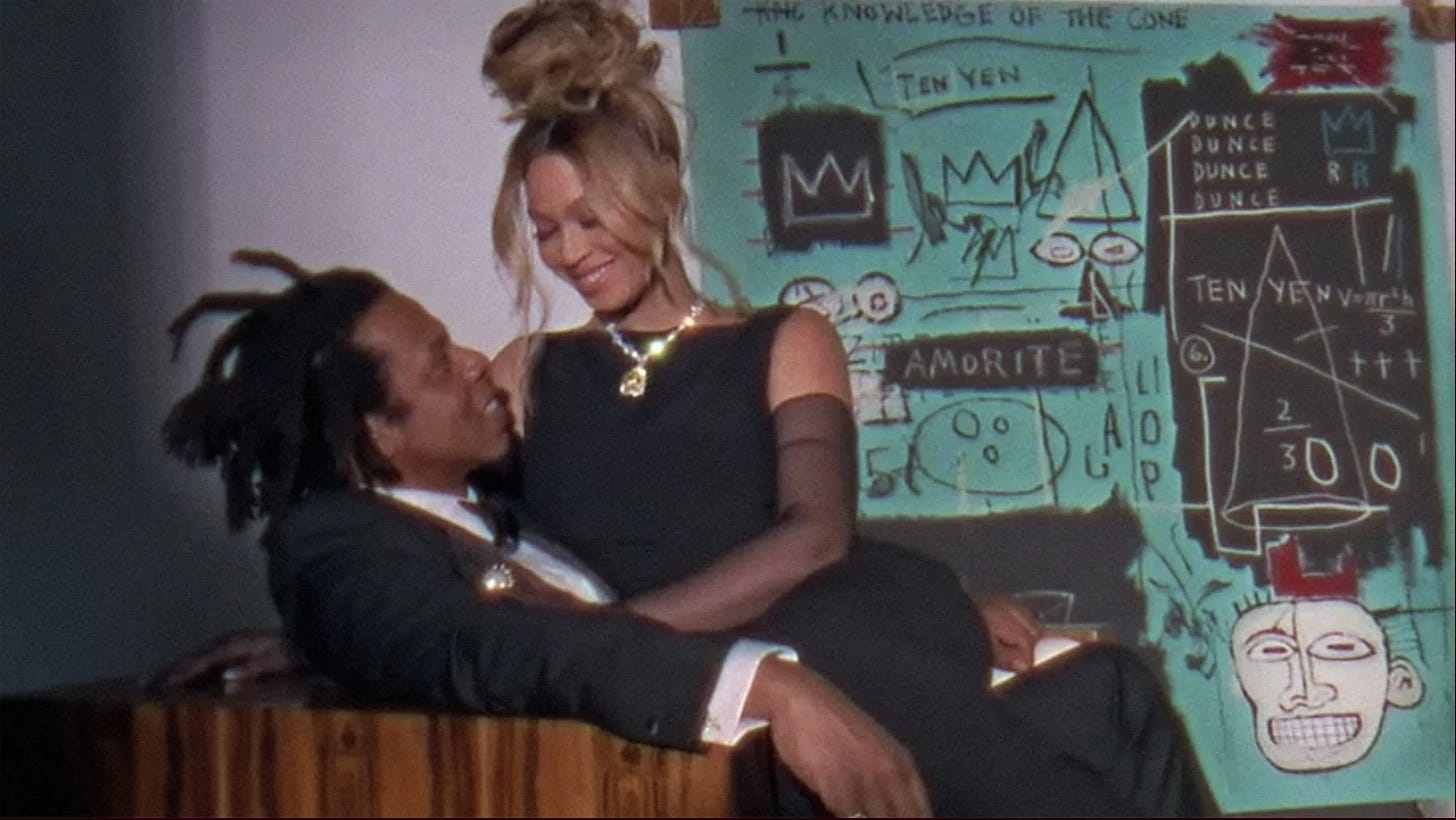 a still from Tiffany and Co. Diamond’s controversial ad with Jay-Z and Beyoncé: Beyoncé, wearing a yellow blood diamond around her neck, sits on her husband’s lap. They gaze at each other smiling softly and clad in all black formal wear. In the background is a work of art by Jean-Michel Basquiat.