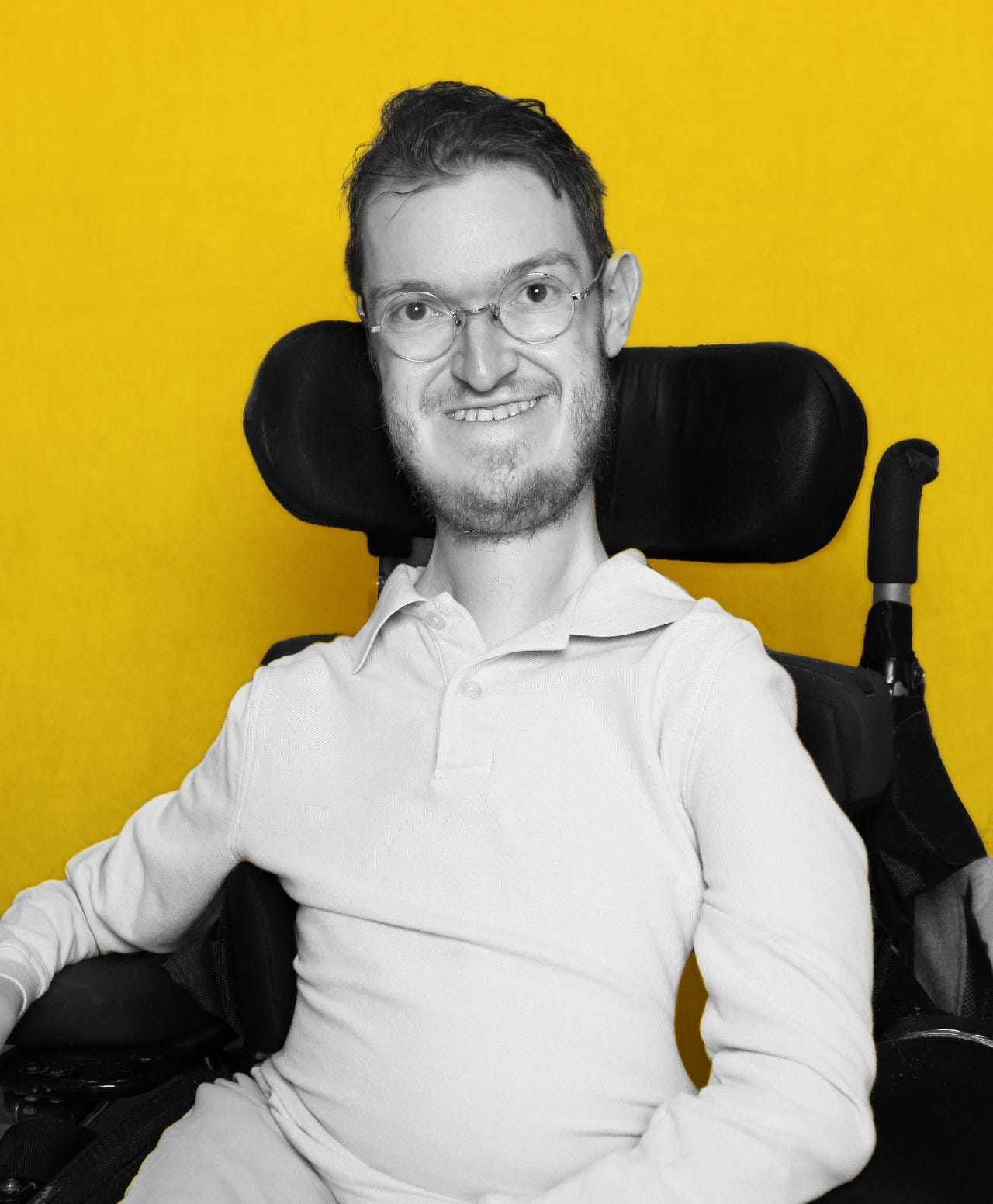 Steven is a white man with short brown hair who wears round, clear glasses. He sits in an electric wheelchair in front of a yellow background. He is wearing a white polo shirt and is smiling at the camera. Steven is a white man with short brown hair who wears round, clear glasses. He sits in an electric wheelchair in front of a yellow background. He is wearing a white polo shirt and is smiling at the camera.