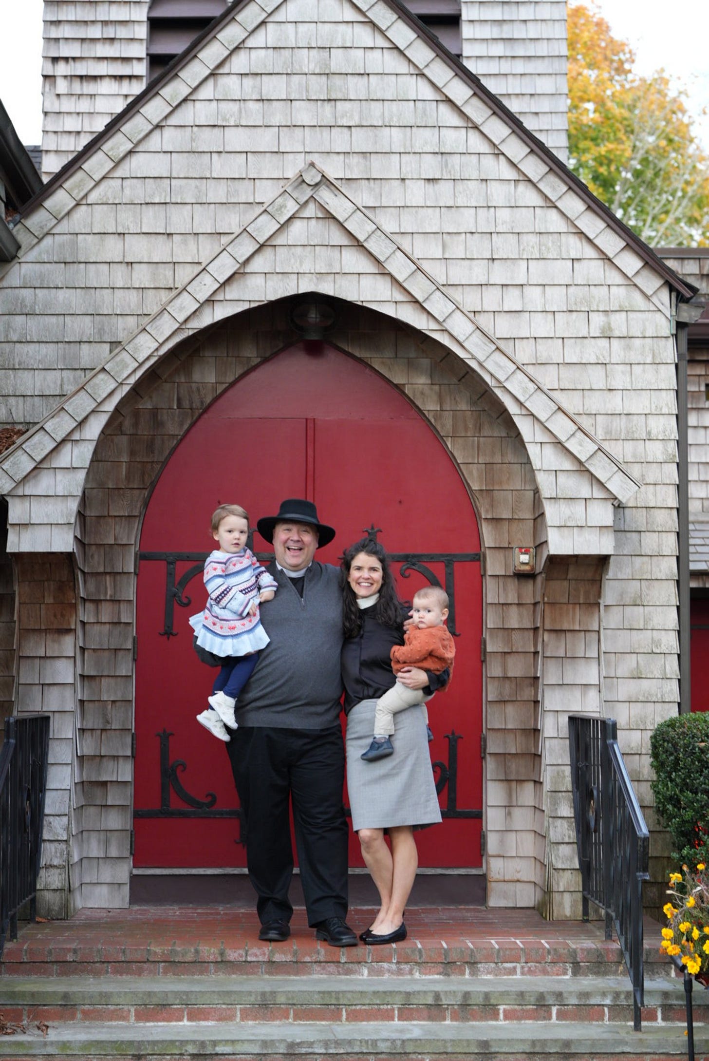Natalie & Thomas, each holding a small child and dressed in their clerical shirts, stand in front of traditional red church doors.