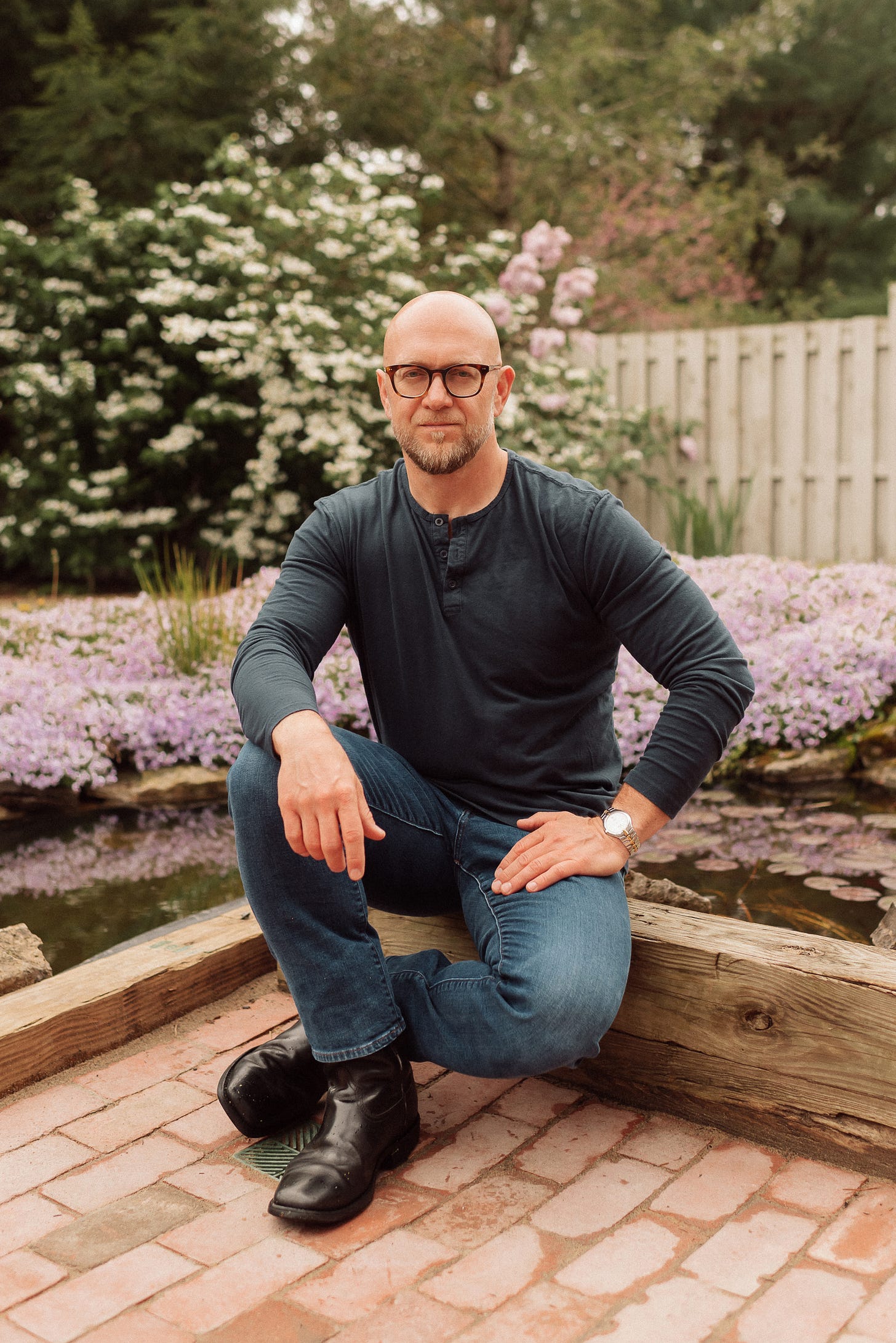 A bald man with a beard and glasses is kneeling on one knee in a garden. He is wearing a dark long-sleeved henley shirt, blue jeans, and black boots. He has a watch on his left wrist and is smiling slightly at the camera. Behind him, there are blooming pink and white flowers, a wooden fence, and lush green foliage, creating a serene and picturesque background.