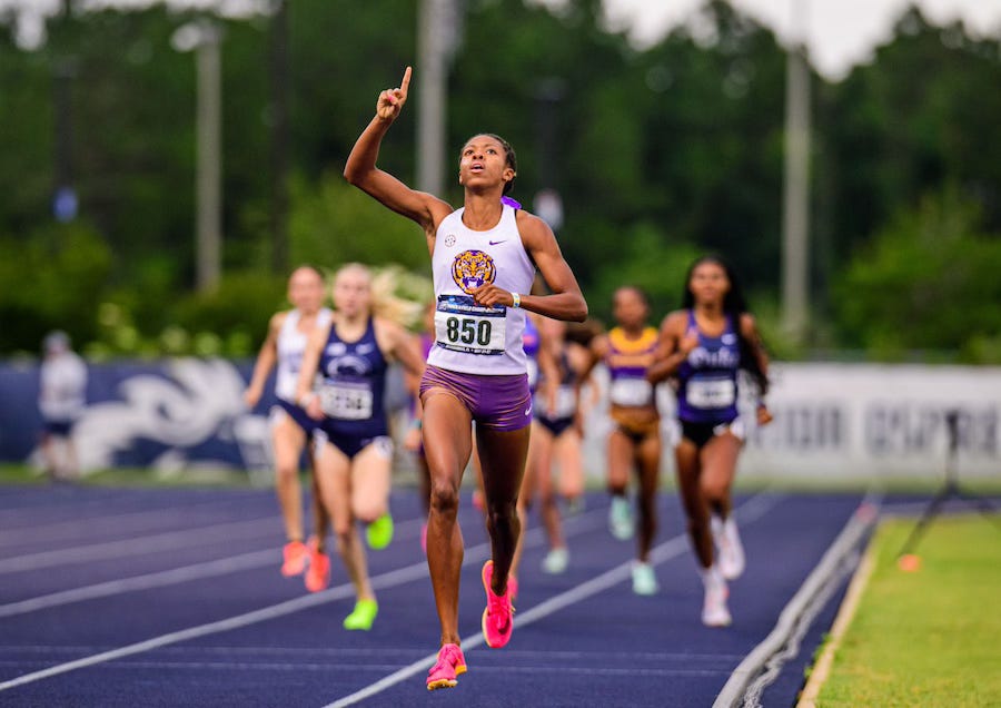 Wearing a LSU uniform with a white top and purple bottoms, Michaela Rose crosses the finish line of a track race with her right arm in the air and one finger pointing toward the sky. There are at least six other runners visible behind her, but she's far enough ahead that they are blurry in the background.