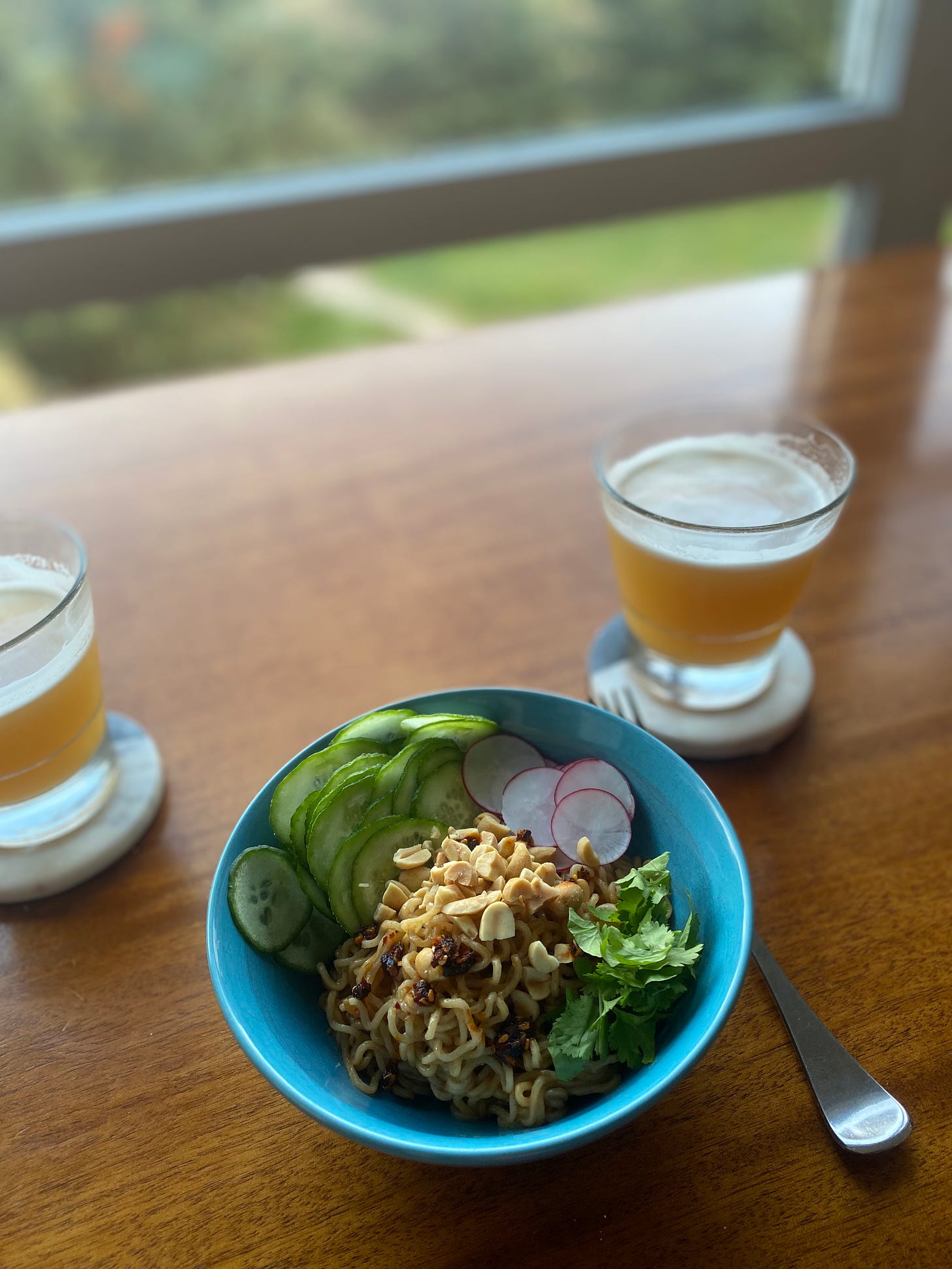 A blue bowl with instant ramen in sesame sauce, and topped with thinly sliced cucumber and radish, along with chopped peanuts, cilantro, and chili crisp. There are glasses of beer on coasters at either edge of the frame.