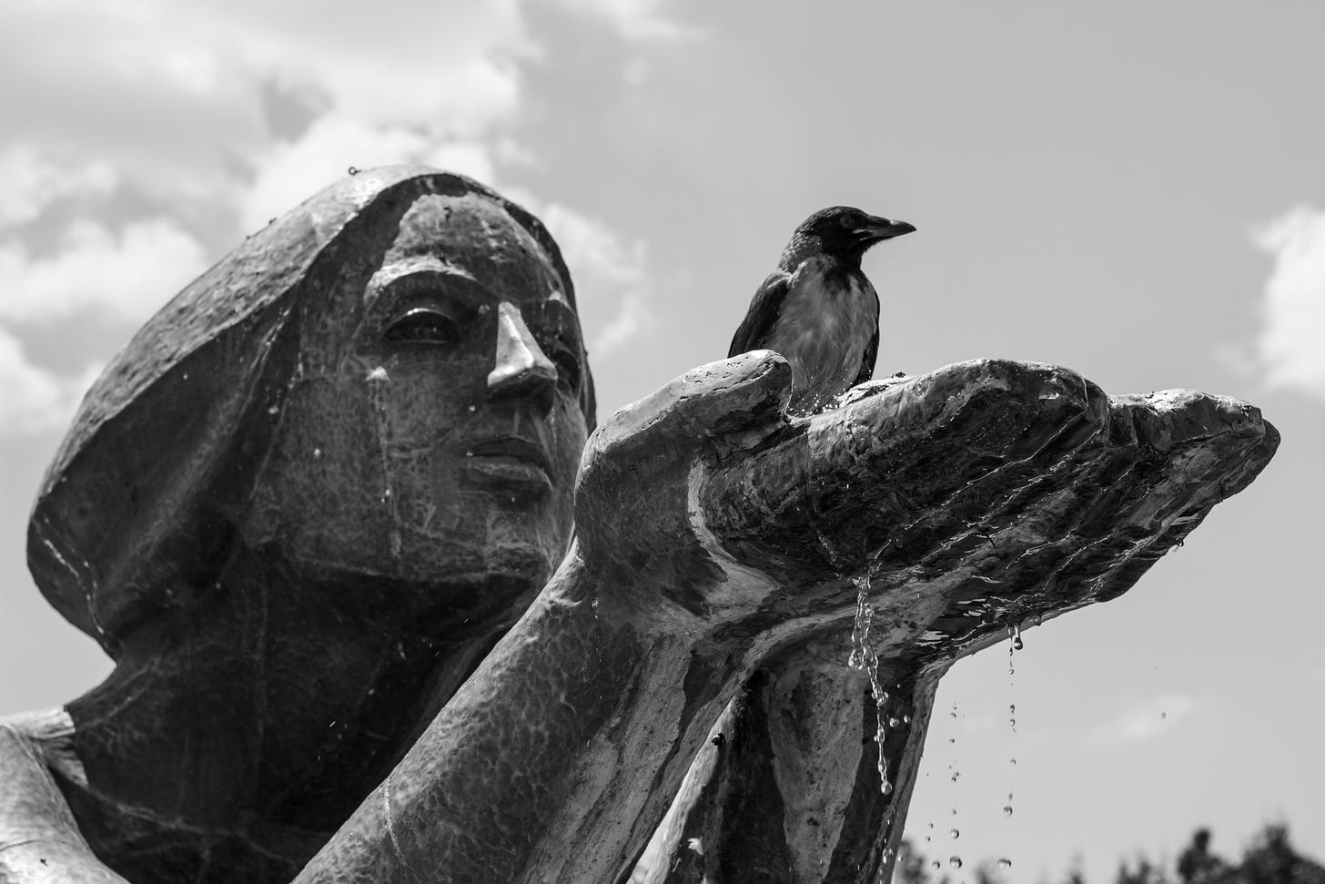 Black and white photo. Low angle view of a fountain statue against the sky. A human figure holds its hands in front of its face, palms to the sky. A bird seats on the hands, and there’s water dripping from the fingers. The statue seems to be looking at the bird nestled in its gentle grasp.