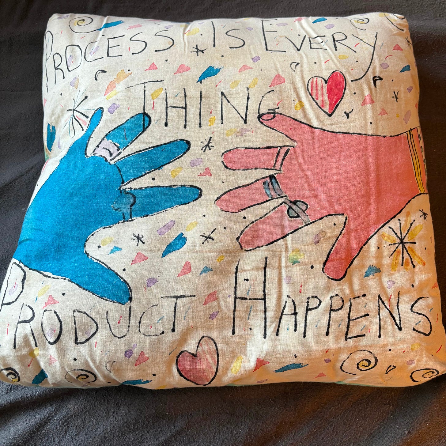 A hand-painted pillow shows one blue hand and one red hand, with hearts and other symbols in the background. Words read "Process is everything, product happens."