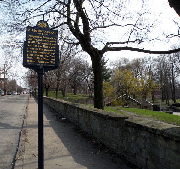 The low stone wall is seen running along the 40th Street side of Arsenal Park with the Pennsylvania Historical Marker for the Allegheny Arsenal to the left and, on the right behind some trees, a portion of the low-lying stone powder magazine is visible in the park.