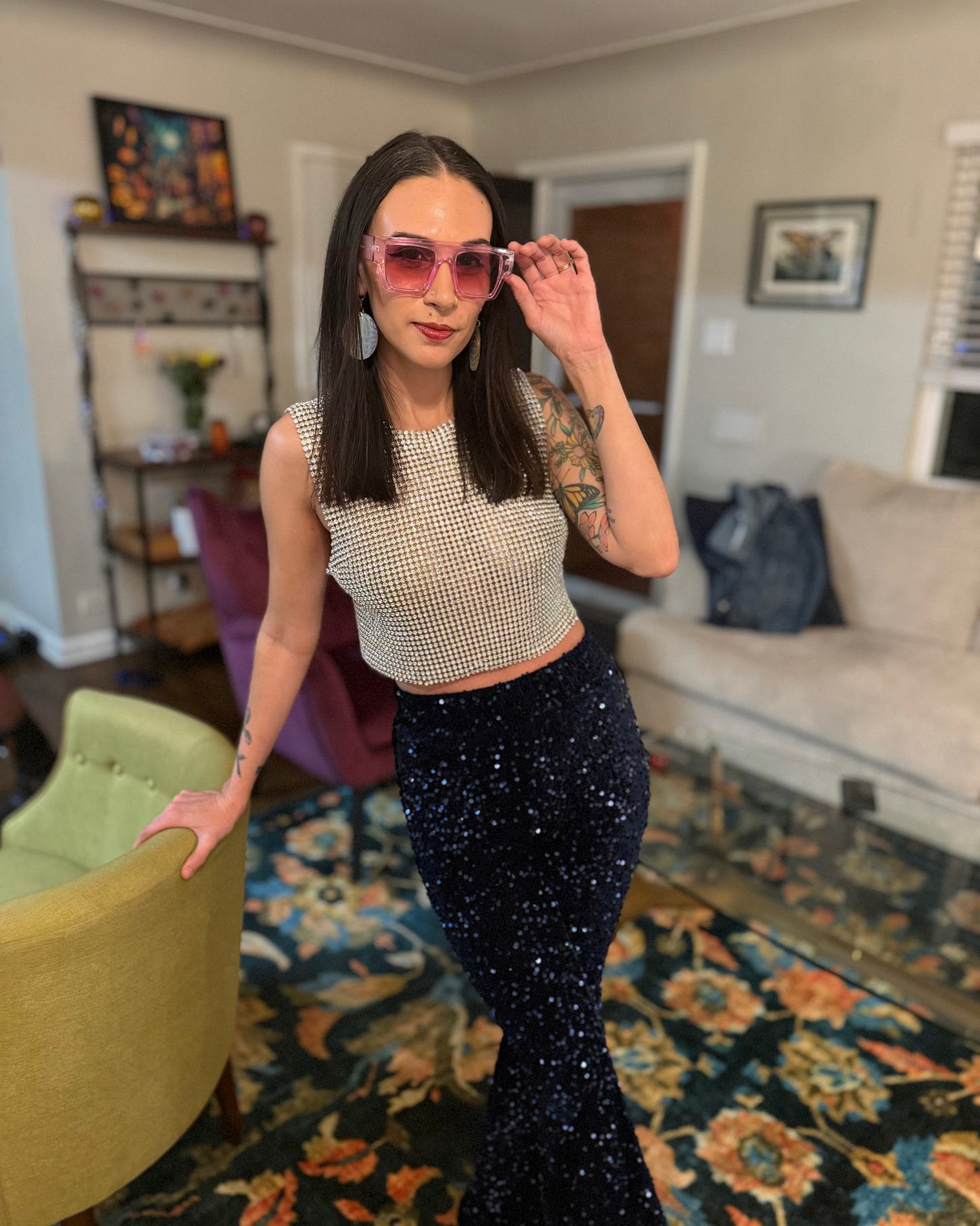 Shohreh is resting her hand on an armchair and wearing glittery blue flare disco pants, a sparkly silver chainmail top, and light purple sunglasses, with long straightened brown hair