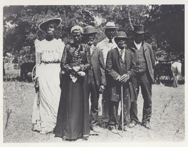 A group of people in fancy dress from the turn of the 19th century, standing together outside. A group of people in fancy dress from the turn of the 19th century, standing together outside.