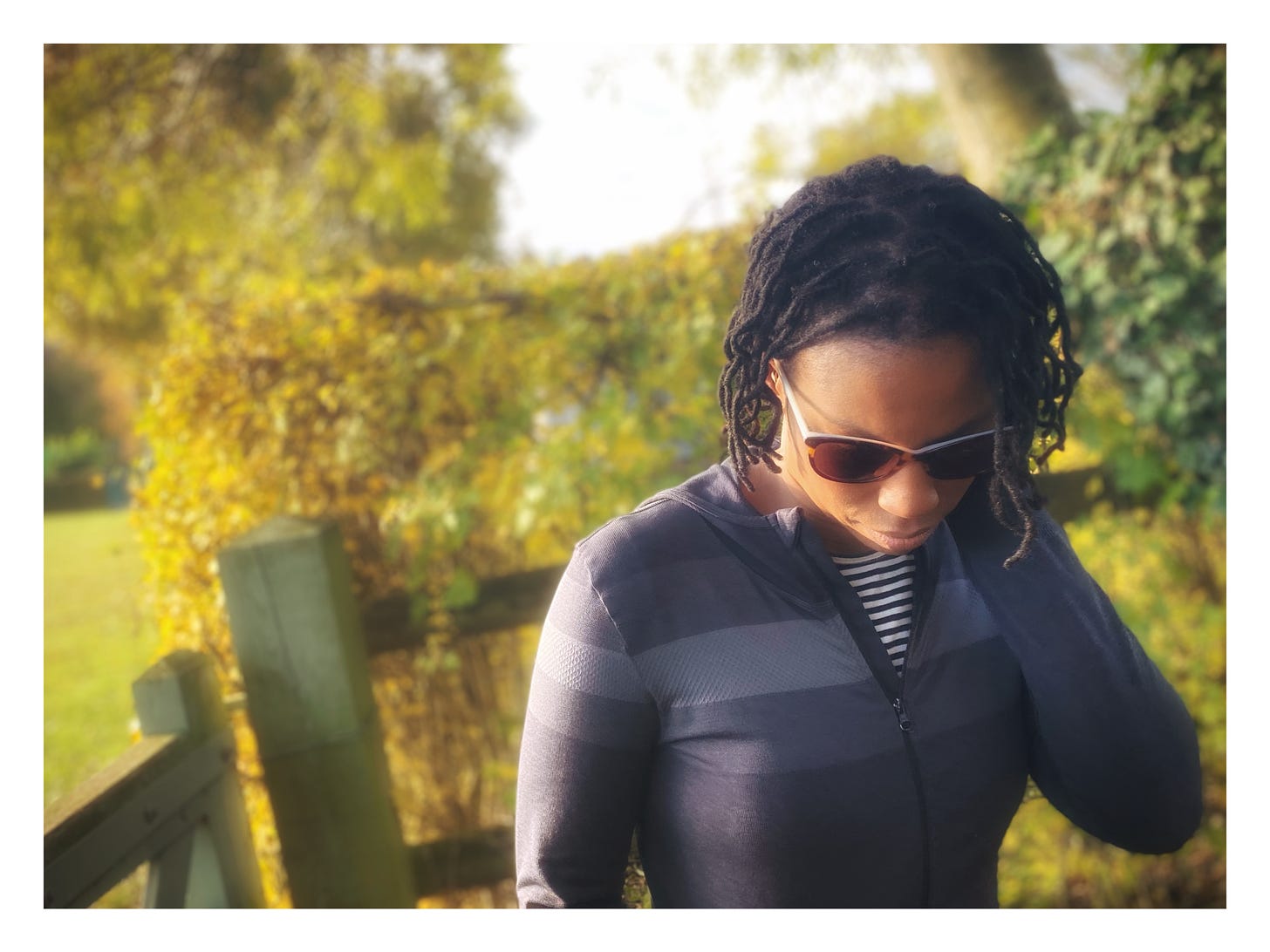 Sandra wears sunglasses, head slightly down cast with Black locs wearing a grey longsleeved shirt with a yellow leaves and fence in background