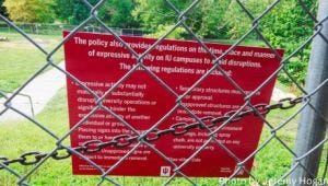 A red sign listing rules about the time, place, and manner of expressive activity appears with a metal chain and a chain-link fence surrounding an Indiana University campus meadow.