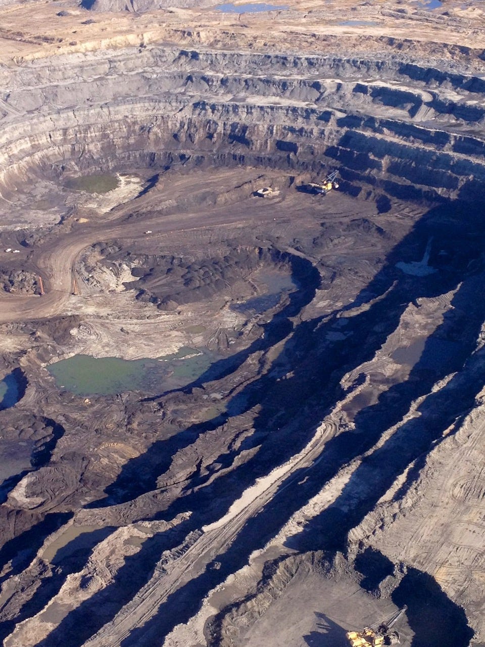 A large open-pin bitumen mine in northern Alberta, showing large industrial equipment as mere specks against the size of the pit.
