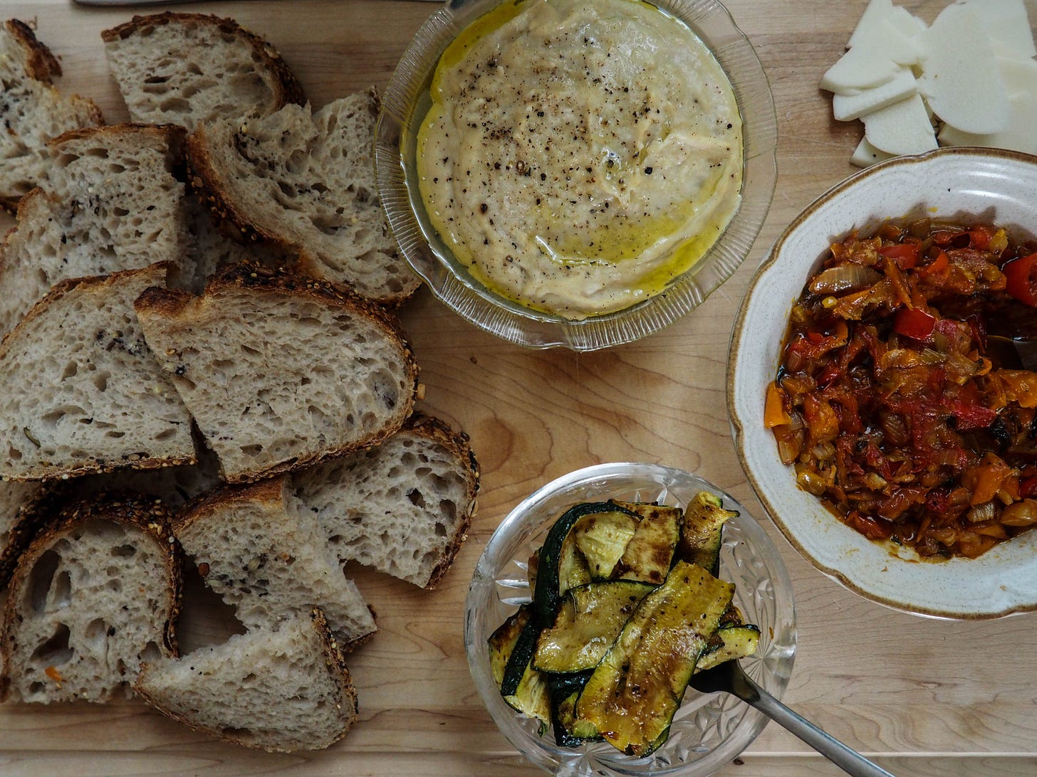 A spread on a cutting board. Bread to the left. In the centre there are two bowls. On top is a bowl with the white bean dip which is beige in colour and drizzled with olive oil. On the bottom is a bowl of grilled zucchinis. To the right is the final bowl which is filled with a red pepper dip. Above the red pepper dip are a few slices of a hard white began cheese.