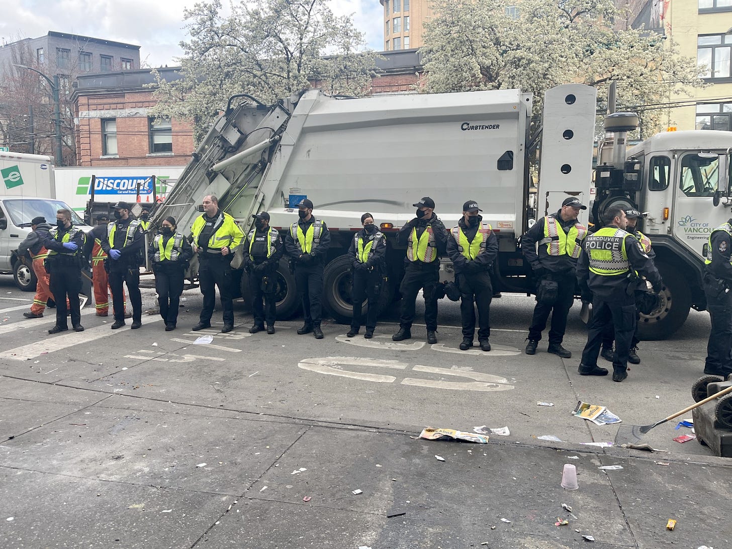 A line of police officers stand beside a garbage truck during a decampment sweep of Hastings Street in April 2023.
