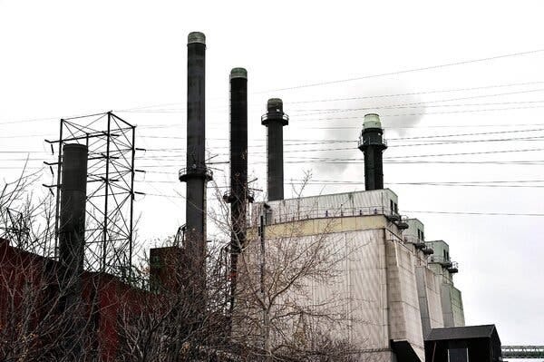 A boxy, grayish structure with tall stacks on a gray winter day. There are leafless trees in the foreground and power lines in the near background. 