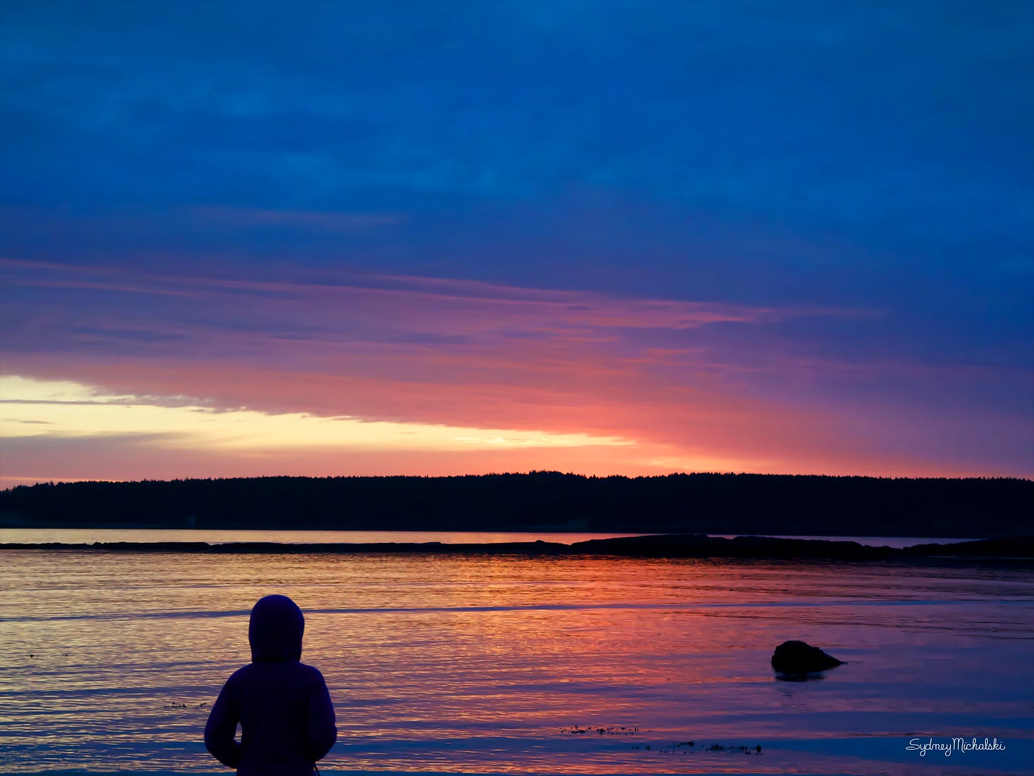 The silhouette of a child gazes out across a calm ocean reflecting a warm sunrise.