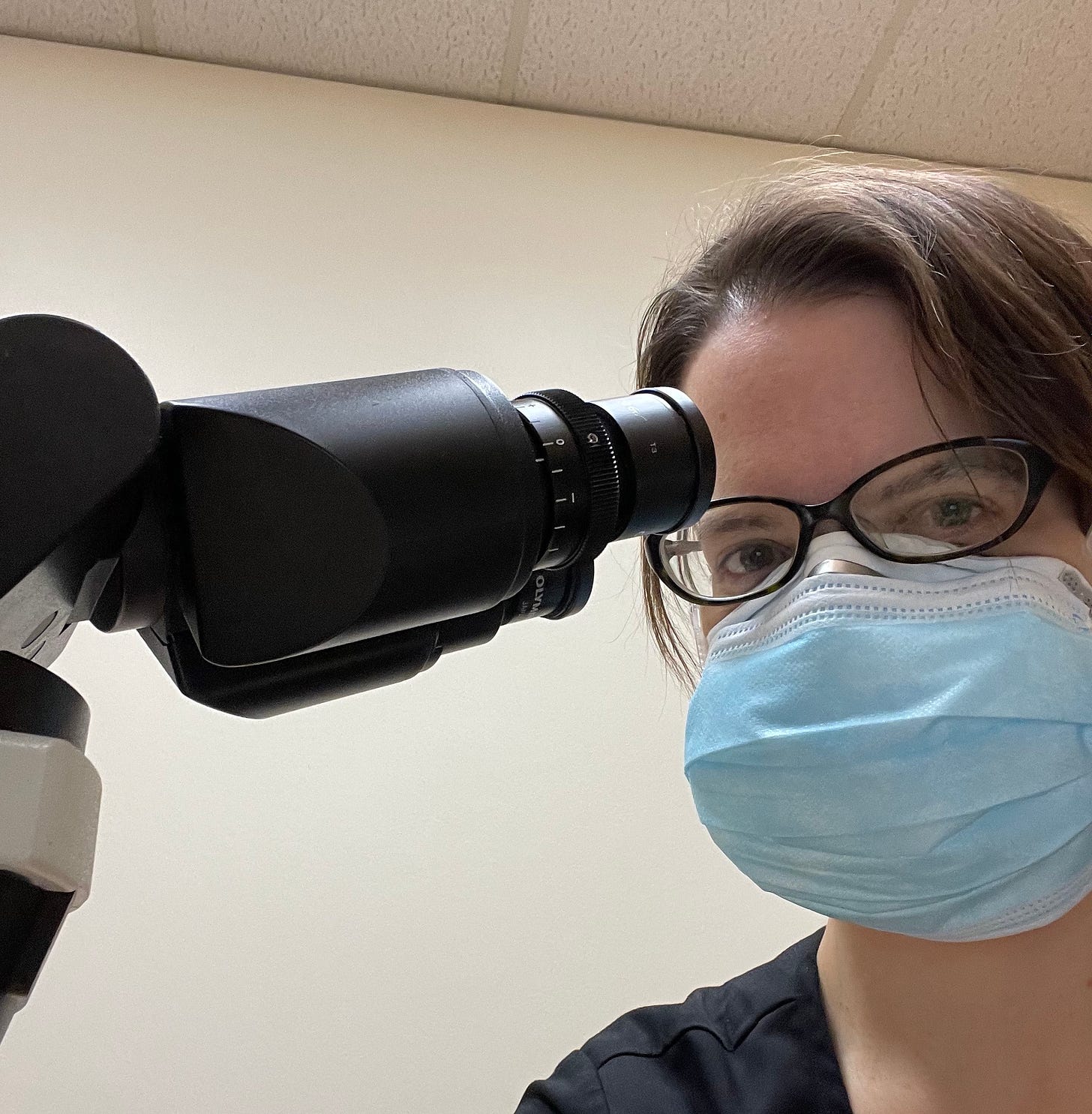 A woman wearing glasses and double masking with a respirator and surgical mask sits in front of a microscope.