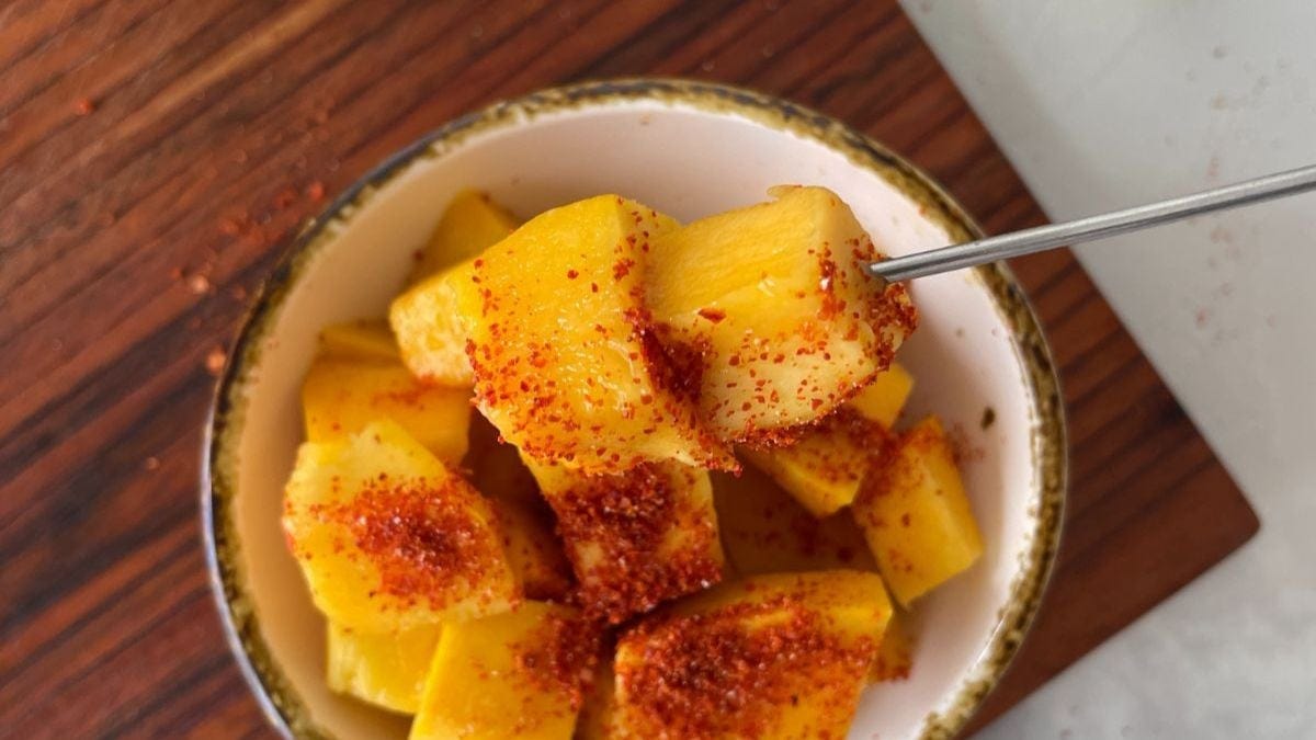 A bowl of freshly cut mango chunks sprinkled with red chili powder, viewed from above on a wooden surface. a spoon is partially inserted into the mangoes.