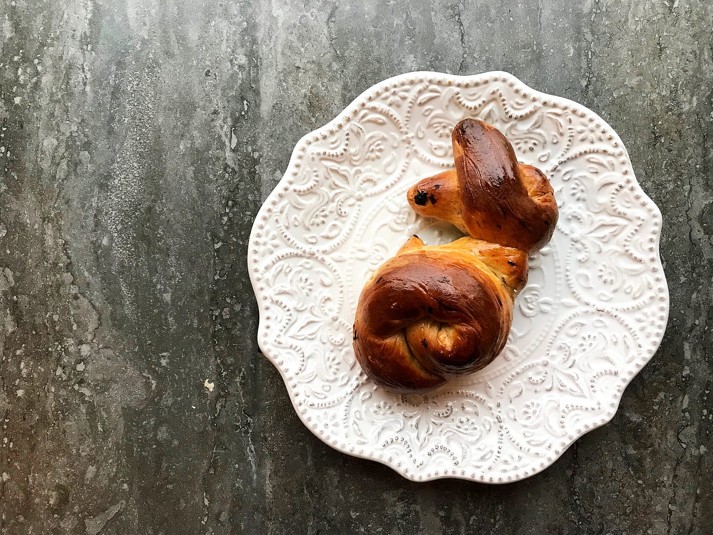 A baked hot cross bunny on an embossed white plate. You can see the twists of dough in shades of gold and sienna. There's a bit of currant on one ear, making it look as if it has a spot. The glaze is shiny. It looks as if it's hopping away. 