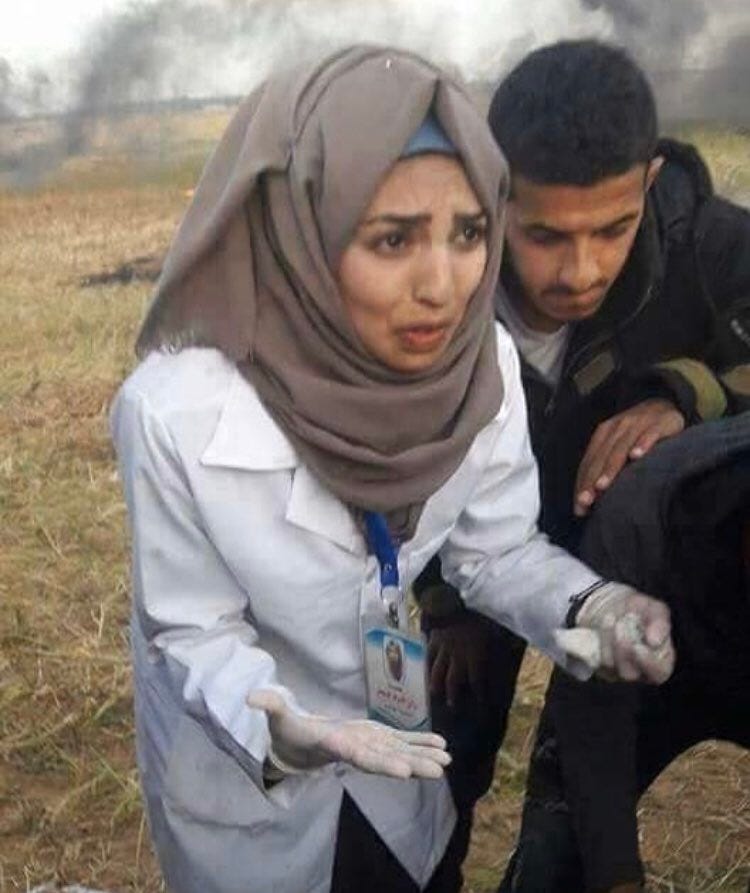 Rouzan al-Najjar in a hijab and white coat pleads with someone off-screen. She wears her medical badge and a blue lanyard around her neck. A male comrade dressed in black is behind her. 